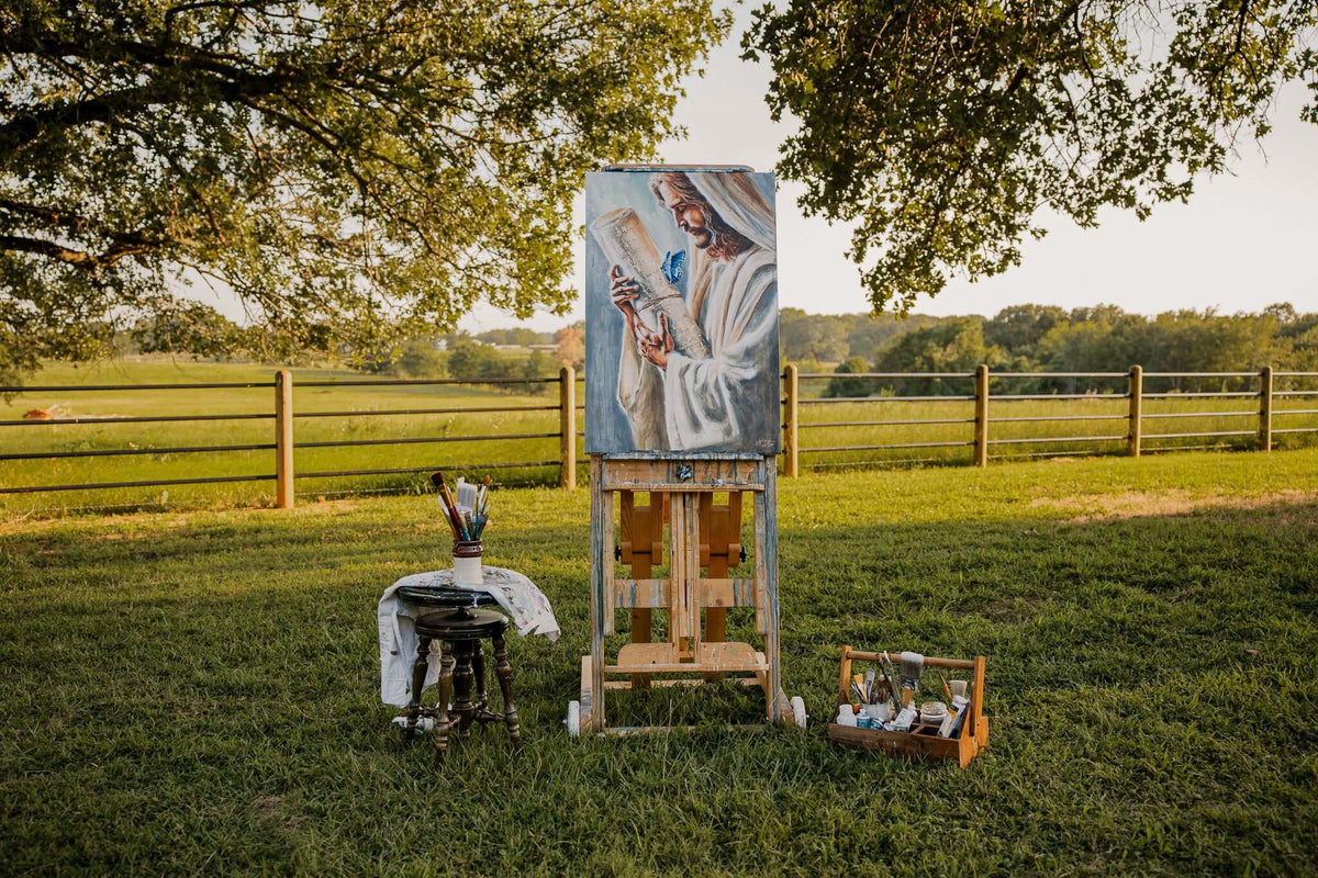 

A Jesus painting displayed on an easel outdoors beneath a tree.